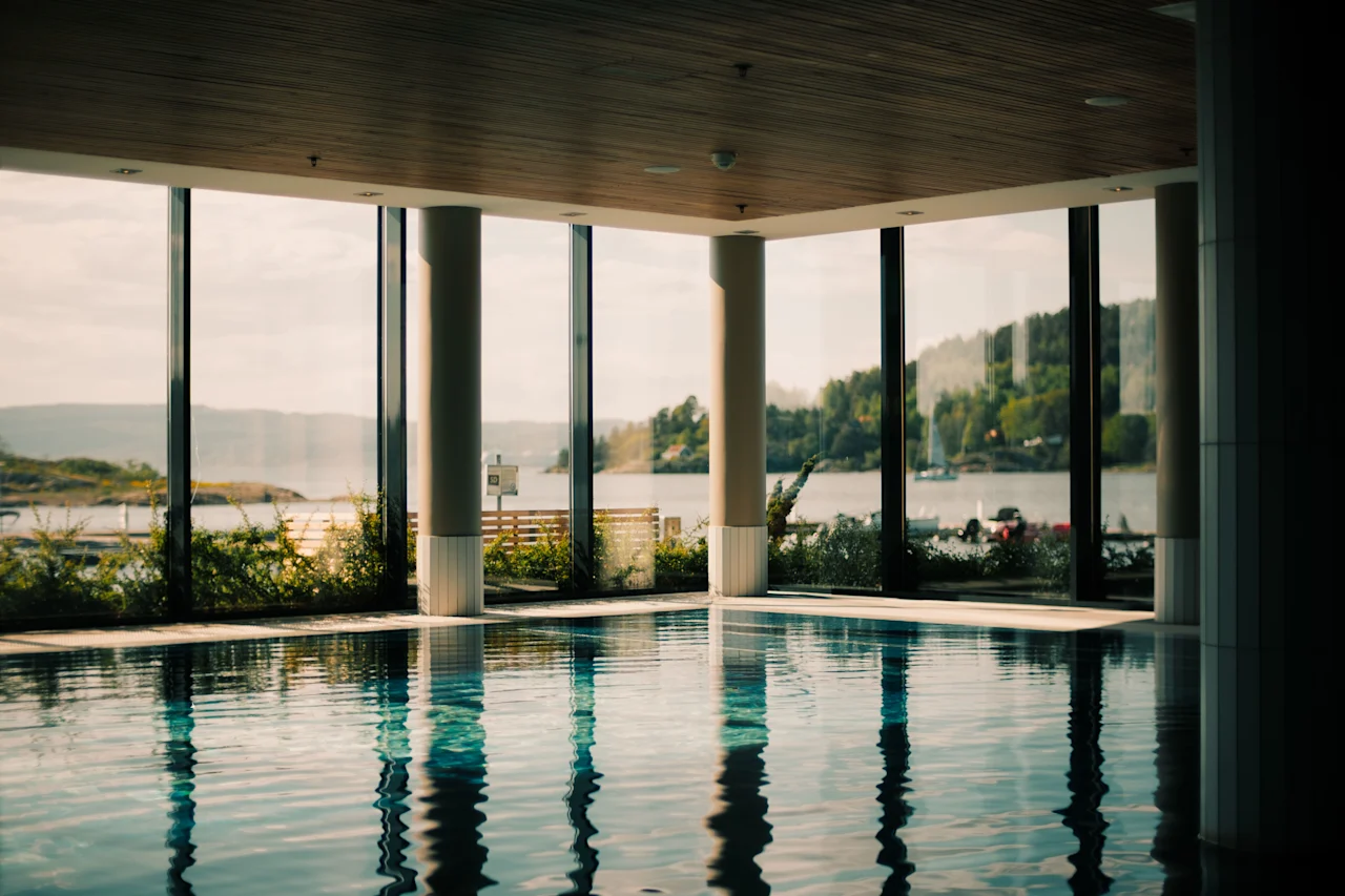 En indendørs swimmingpool med store vinduer, der tilbyder en naturskøn udsigt over en sø omgivet af frodige grønne bakker under en lys himmel.
