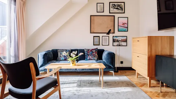A blue velvet sofa adorned with colorful pillows sits in a living room, facing a wooden coffee table with a potted plant, amidst framed artwork and a wooden cabinet.
