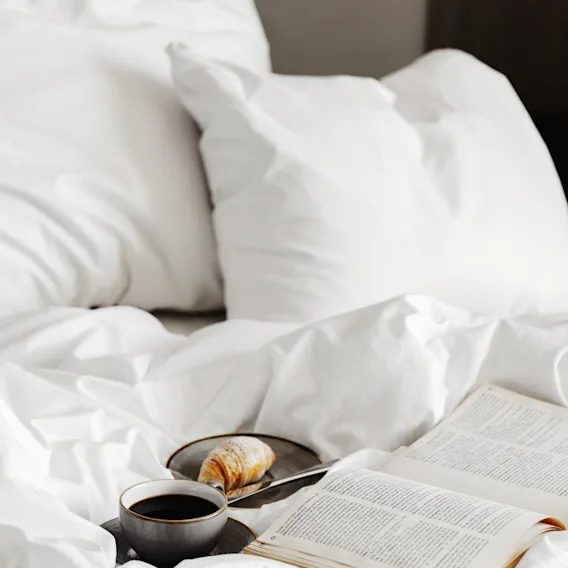 A cozy bed with white linens, an open book, a cup of coffee, and a croissant, suggesting a relaxing morning.