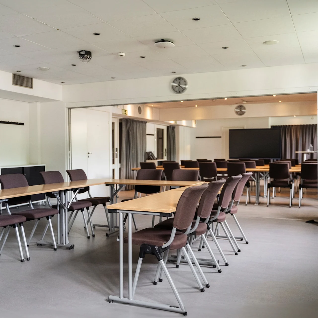 Bord som är ordnade i en U-form med stolar i ett rymligt, ljust konferensrum. Vita väggar, en klocka och en nödutgångsskylt. Gardiner täcker delvis en stor skärm.