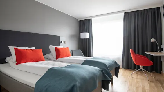 Två prydligt bäddade enkelsängar med röda kuddar och blå täcken i ett hotellrum. En röd stol och ett skrivbord står vid ett stort fönster med mörka gardiner.