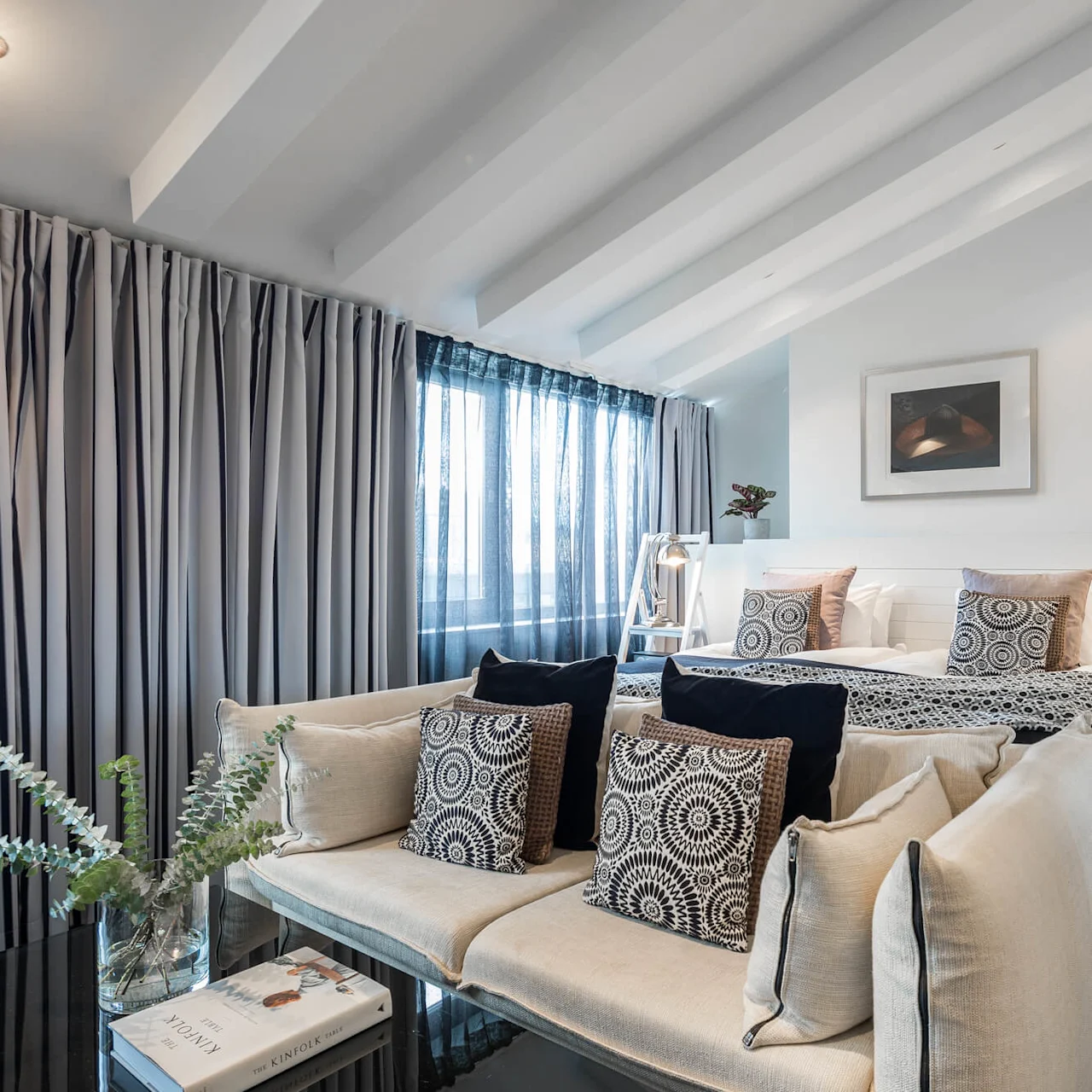 Et moderne soverom har en seng med mønstrede puter, en koselig sofa og store vinduer med gardiner. En speilvegg og et sidebord med lampe tilfører eleganse.