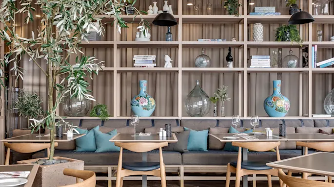 Wooden tables and cushioned benches in a cozy dining area, warmly lit, with decorative shelves and greenery.