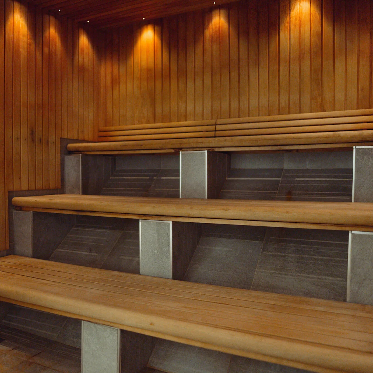 En sauna med træbeklædning byder på niveaudelte bænke, hvilket skaber en varm og indbydende atmosfære. Blød belysning øger den hyggelige stemning.