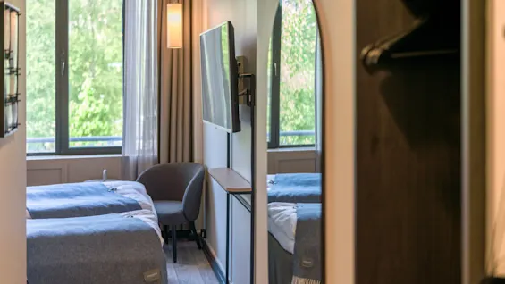A modern, neatly arranged hotel room features twin beds and a wall-mounted TV. Soft lighting and a large window illuminate the serene space, complemented by a chair and light-colored decor.