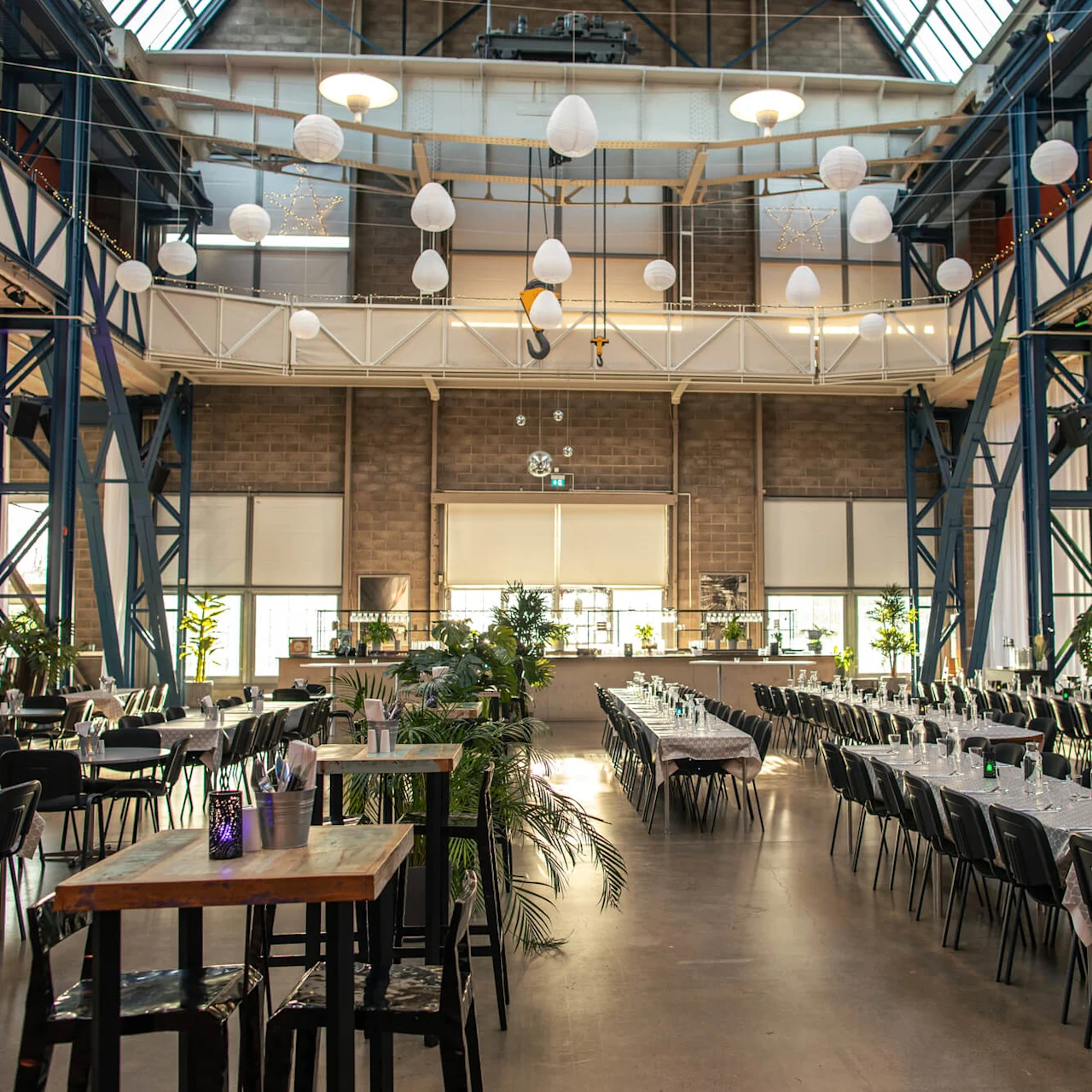 Tables are set with chairs and cutlery, arranged in rows under hanging lights in a large, industrial-style hall with high ceilings and plants.