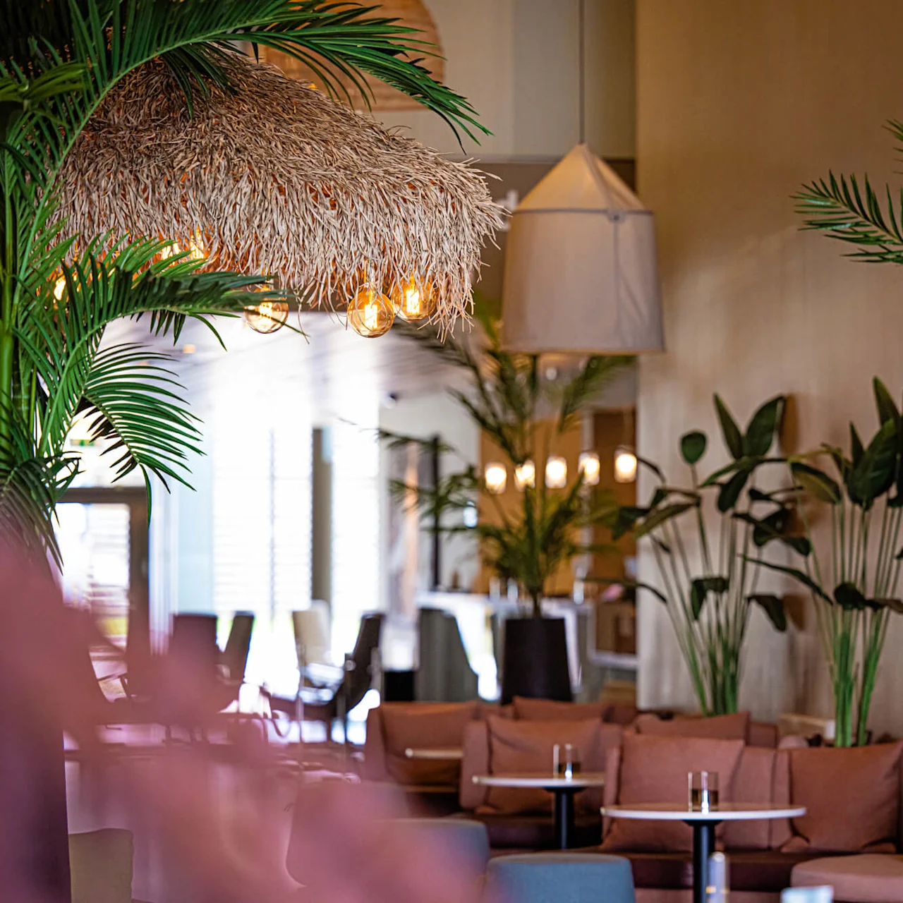 Myke sitteplasser er arrangert i en varmt opplyst lounge pyntet med tropiske planter og en taklampe i stråtak. Runde bord med drikkevarer skaper en koselig, innbydende atmosfære.