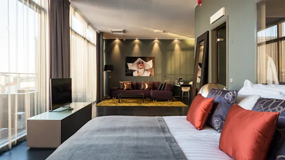 A luxurious bedroom features a large bed with patterned pillows, adjacent to a sitting area with a couch and art on the wall, surrounded by tall windows and soft lighting.