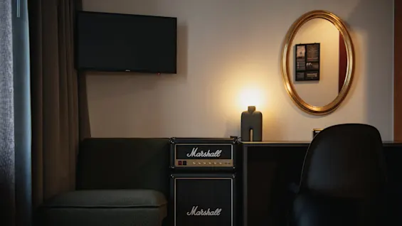 En Marshall-förstärkare står på en mörk soffa under en väggmonterad TV. I närheten lyser en lampa upp en rundad spegel och en svart stol mot en ljus vägg.