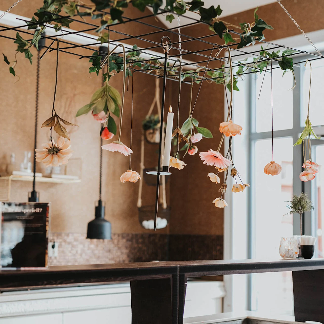 Omvendte blomster hænger fra et loftgitter i en hyggelig café med naturligt lys.