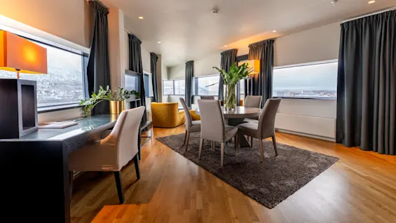 Et spisebord med stole og en vase med grønne planter står på et brunt tæppe i et moderne rum med store vinduer, gardiner og udsigt over sneklædte bjerge.