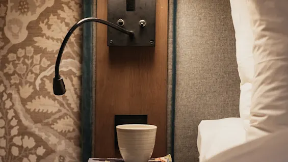 En mysig sängscen med varm lampa, läslampa, kopp och böcker på nattduksbord bredvid säng. Synlig text: ROSAMUNDE PILCHER Forste sommer, CICERO, HOLT.