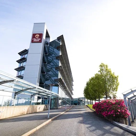 Et høyhus med et "Clarion Hotel"-skilt står i et moderne, beplantet område med grøntområder og blomster, ved siden av en glassgang under en klar himmel.