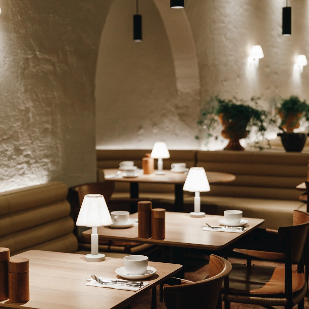 Wooden tables set with cups and saucers are arranged under soft white lamps in a cozy dining area with arched, white-plastered walls and potted plants.