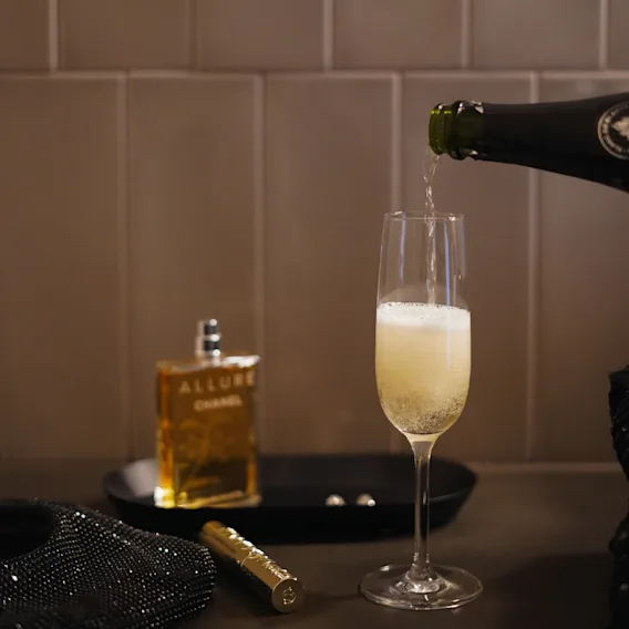 Pouring sparkling wine into a glass next to perfume and accessories, getting ready for a night out.