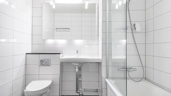 Et godt opplyst, moderne bad med hvite flislagte vegger, et toalett, vask og et badekar med glassdusjskjerm.