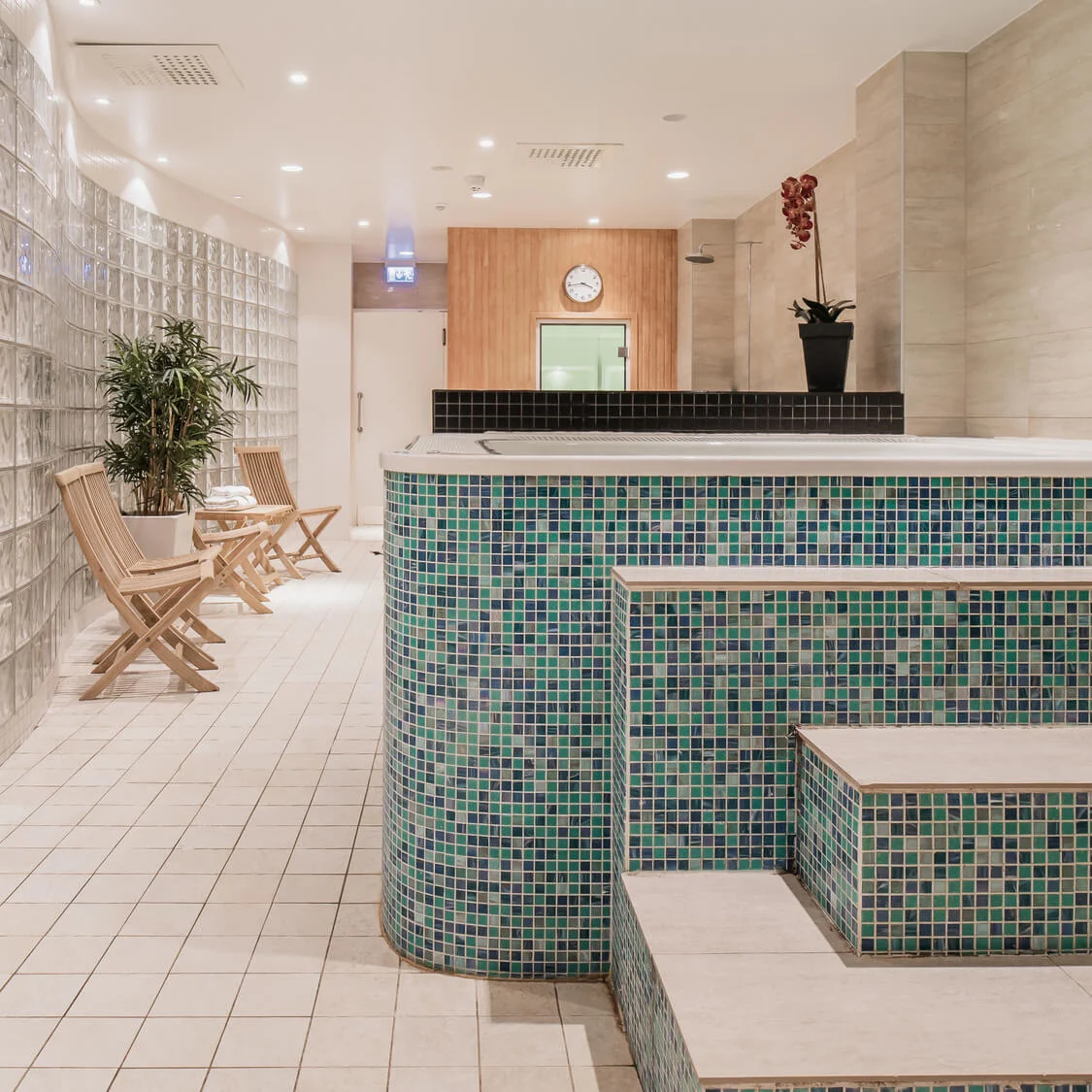 A tiled jacuzzi sits in a bright spa room, surrounded by glass block walls, potted plants, and wooden chairs. A clock hangs on the wooden wall in the background.