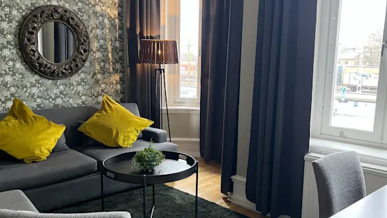 A cozy living room with modern decor, featuring a gray sofa with yellow pillows, and large windows draped in dark curtains.