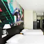 Two beds with white linens are located in a modern hotel room. A colorful DJ mural is on the wall, and a hanger and shelf are near the entrance.