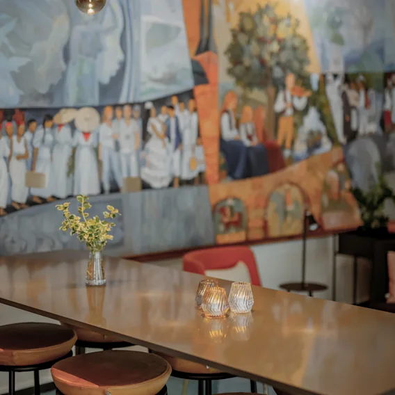 Bord med afføring under gyldne vedhængslamper i et rum med et stort, farverigt vægmaleri, der viser mennesker og naturscener. En lille vase med blomster og stearinlys pynter bordet.