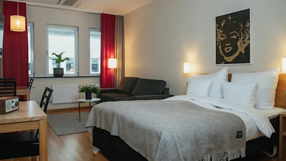 A bed with a gray blanket is positioned in a modern room with wooden flooring. Nearby, a black sofa, red curtains, and a plant by the window create a cozy ambiance.
