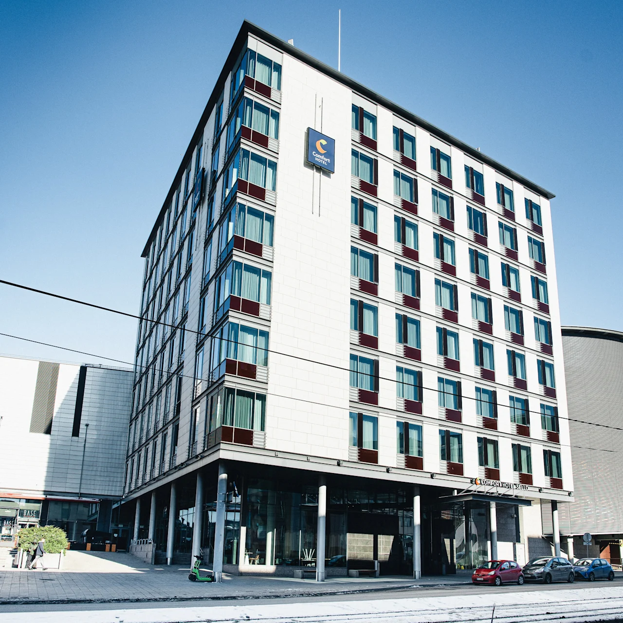 Tall, modern building with glass windows stands on a snowy street. A sign reads "Clarion Hotel Stavanger." Surrounding environment includes blue sky and parked cars.