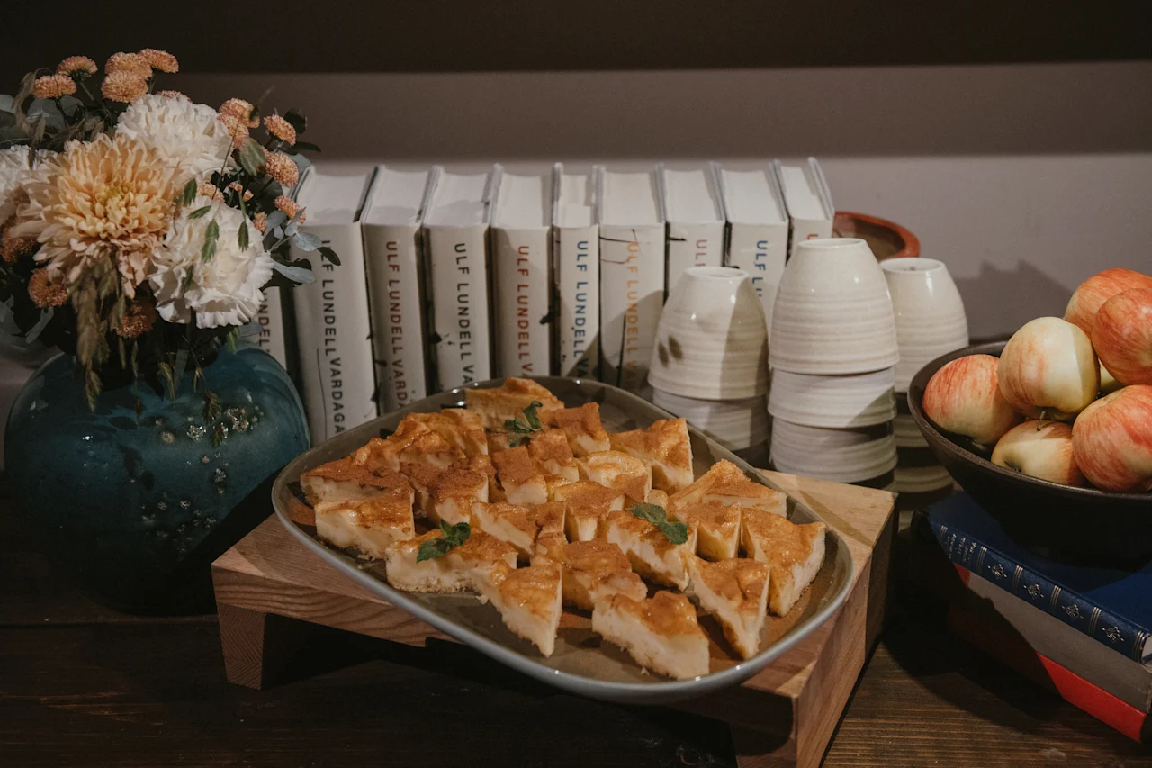 En mysig miljö med en tallrik dessert, staplade koppar, äpplen, blommor och böcker av ULF LUNDELL VARDAGA.