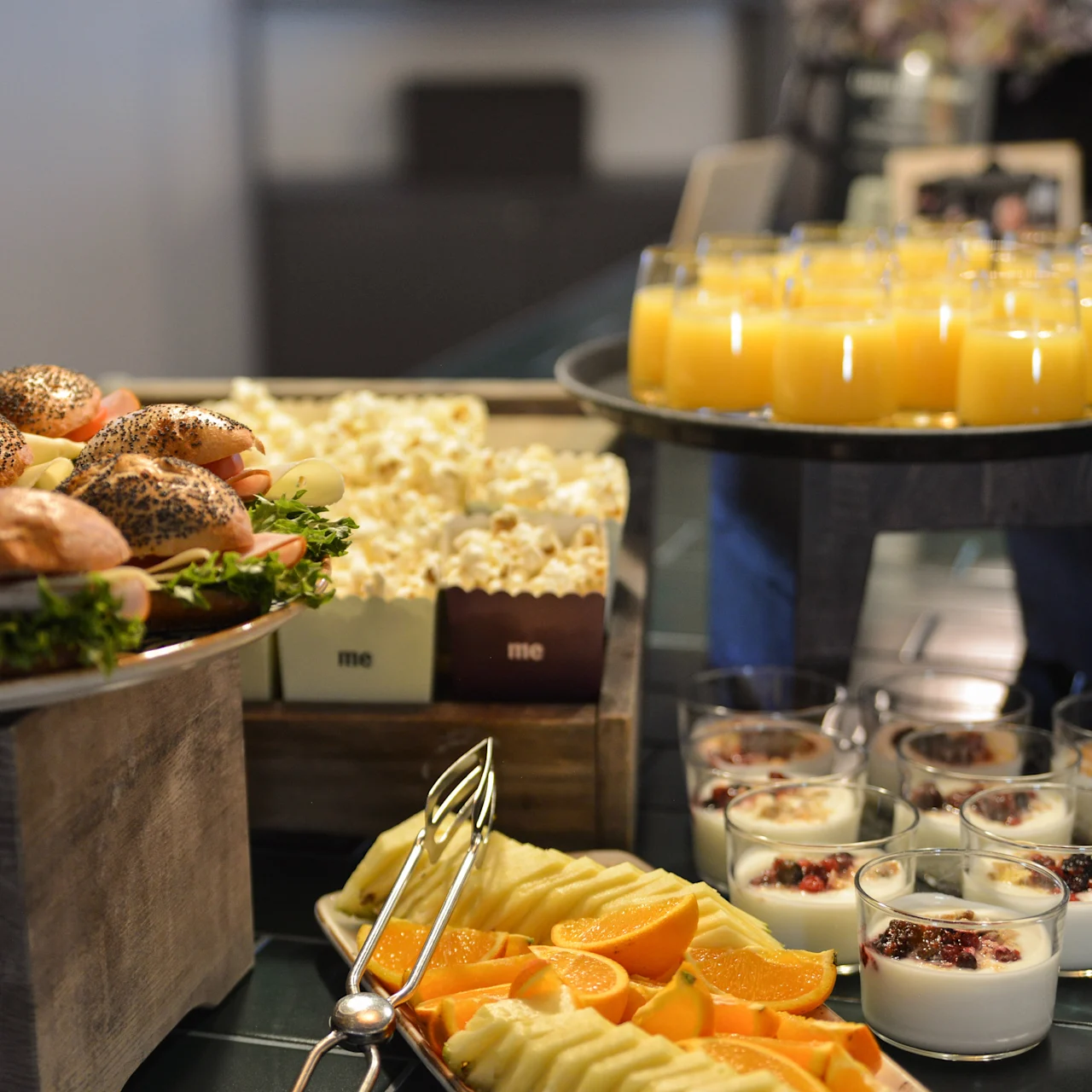 Sandwiches ligger på en træbakke, omgivet af popcorn og yoghurt i glas med frugt. Appelsinjuice er hældt i flere glas på en separat bakke indendørs.