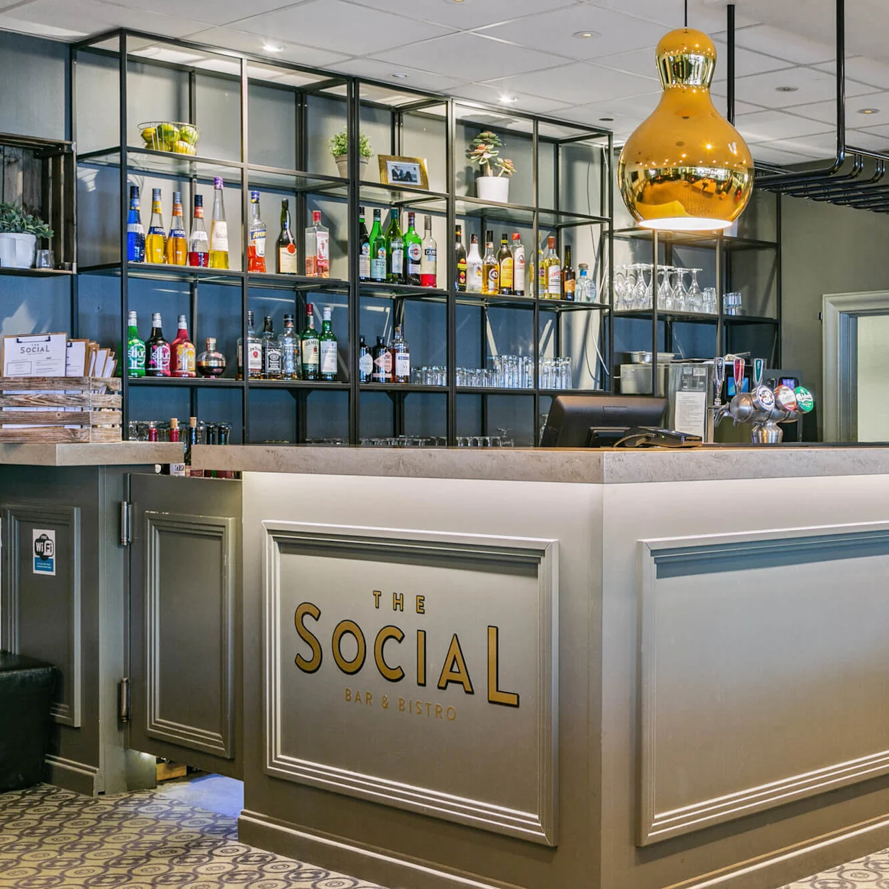 En bar med navnet "The Social Bar & Bistro," hvor flasker og glas er udstillet på hylder. To gyldne pendellamper hænger over disken i en indbydende atmosfære.