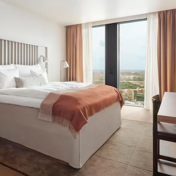 A neatly made bed with white linens and an orange blanket stands by a glass window, providing a view of a cityscape. The room features wooden furniture and beige curtains.