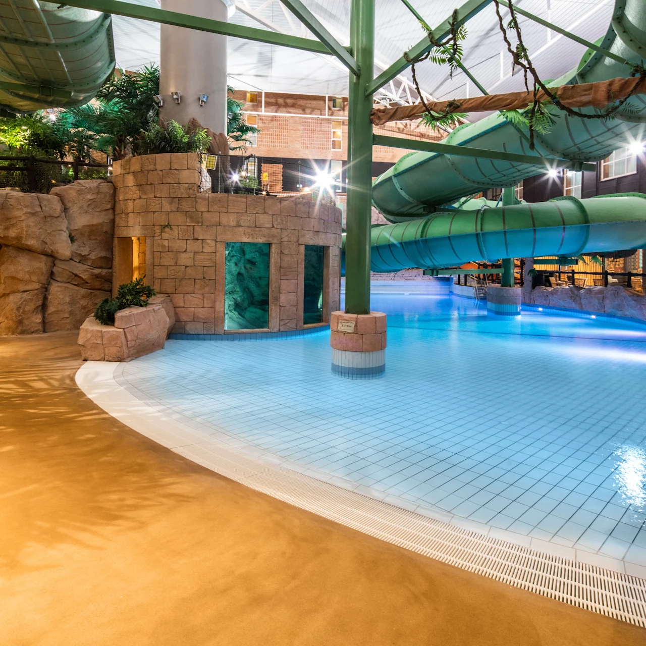 A large green waterslide curves above a clear blue pool in an indoor water park. The area is surrounded by artificial rock formations and plants.