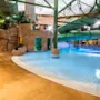 En stor grøn vandrutsjebane bøjer sig over en klarblå pool i en indendørs vandpark. Området er omgivet af kunstige klippeformationer og planter.