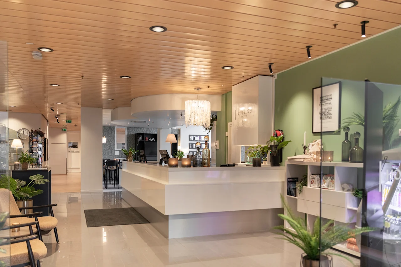 Kutsuva hotellin vastaanottoalue, jossa on pitkä valkoinen tiski, mukavat istuimet ja kasveja. Näkyvä teksti: HOME HOTEL.