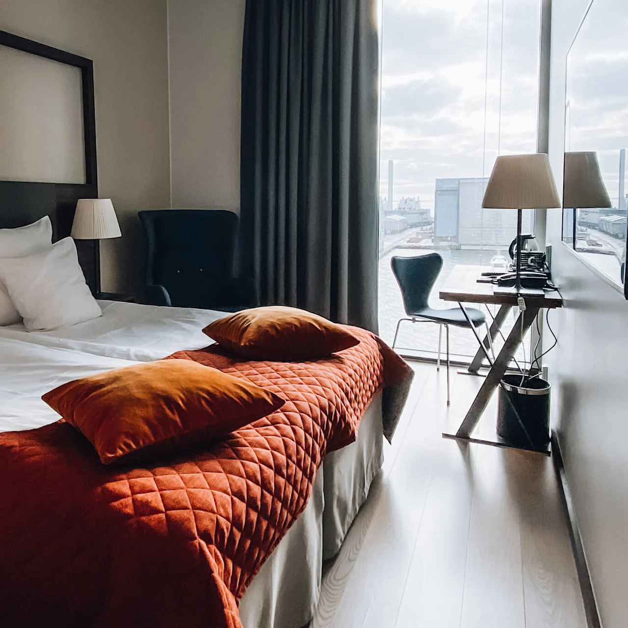 En pænt redt seng med hvidt sengetøj og orange puder står i et moderne hotelværelse med et minimalistisk skrivebord og stol nær et stort vindue med udsigt over en industriel havn.