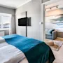 A neatly made bed with teal bedding faces a wall-mounted TV in a modern bedroom. Large windows display a scenic view. Adjacent room has sofa and ocean-themed decor.