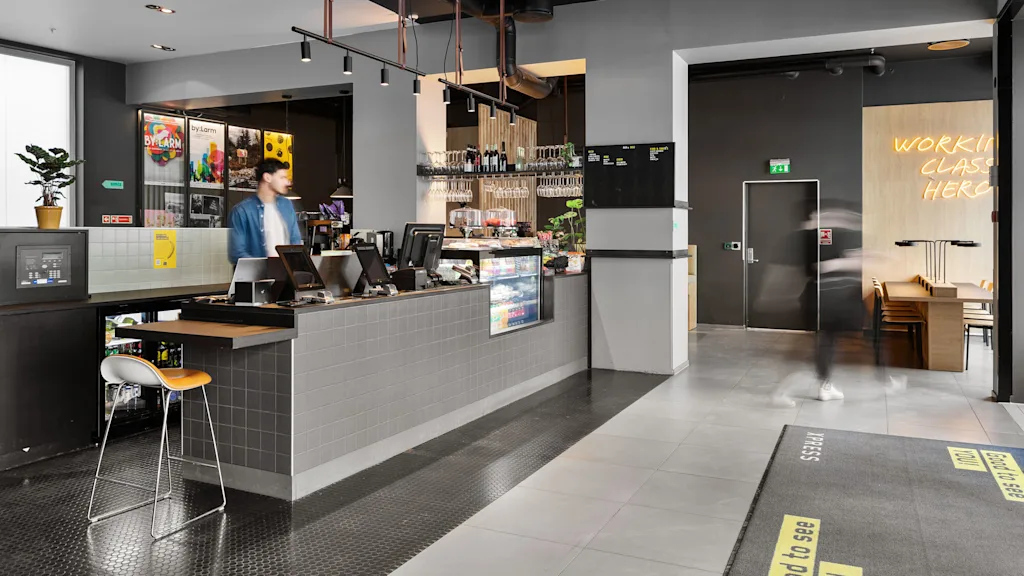 En cafédisk med en person der står, omgivet af moderne indretning og siddepladser. Neonskiltet læser "WORKING CLASS HEROES." Gulvmåtte tekst: "Good to see you" og "Pssss."