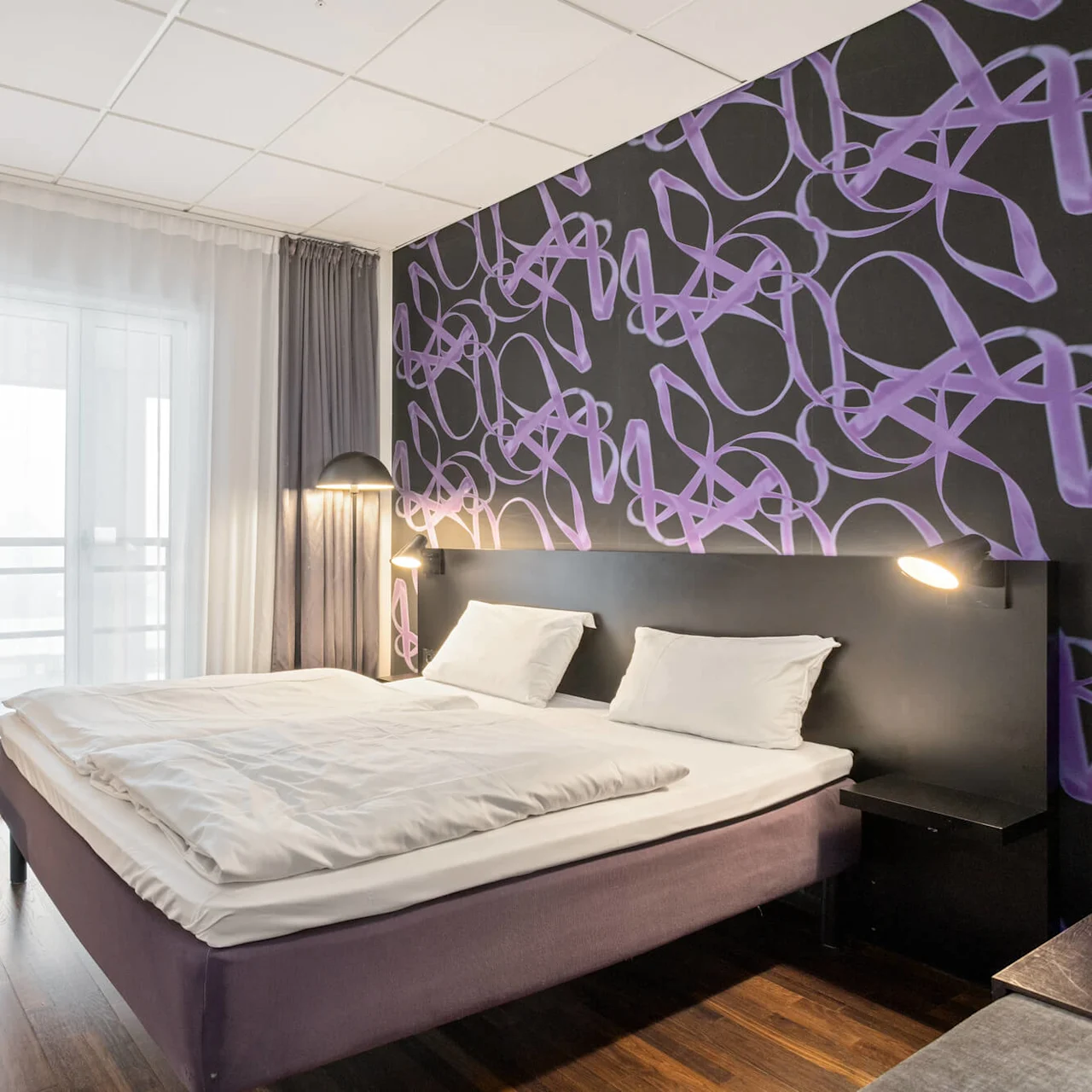 A double bed dominates a modern hotel room, accented with a purple headboard design, white bedding, and wooden flooring.