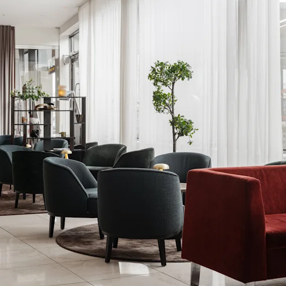 En modern hotellounge med bekväma fåtöljer och röd soffa, upplyst av fönster. 