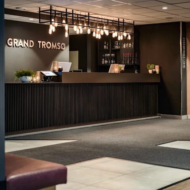 Moderni hotellin vastaanottoalue tummalla puupöydällä, valaistu tyylikkäillä riippuvalaisimilla. Seinällä lukee GRAND TROMSO.