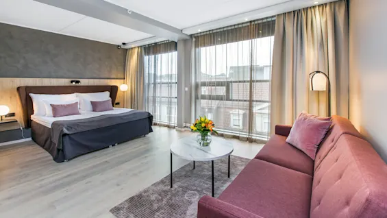 A modern bedroom features a large bed with gray bedding alongside a red sofa, a round table with flowers, and floor-to-ceiling windows with sheer curtains.