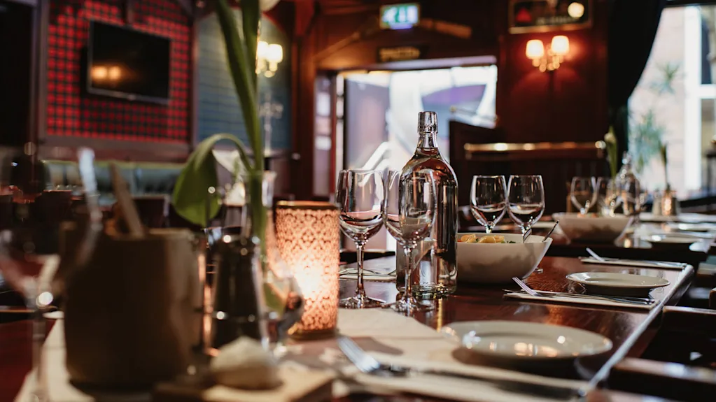Ett bord är elegant dukat med vinglas, tallrikar och ett ljus i en svagt upplyst restaurang. Bakgrunden innehåller mjuk belysning, en TV på en röd vägg och en öppen dörr.