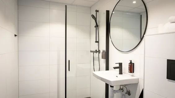 A modern bathroom with a walk-in shower, a sink with a black faucet, and a round mirror. White tiles and dark accents.