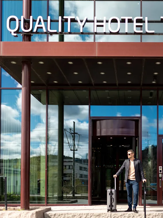 En man med en resväska lämnar ett hotell med glasfasad, under en skylt där det står "QUALITY HOTEL THE WEAVER" en solig dag som reflekteras i fönstren.