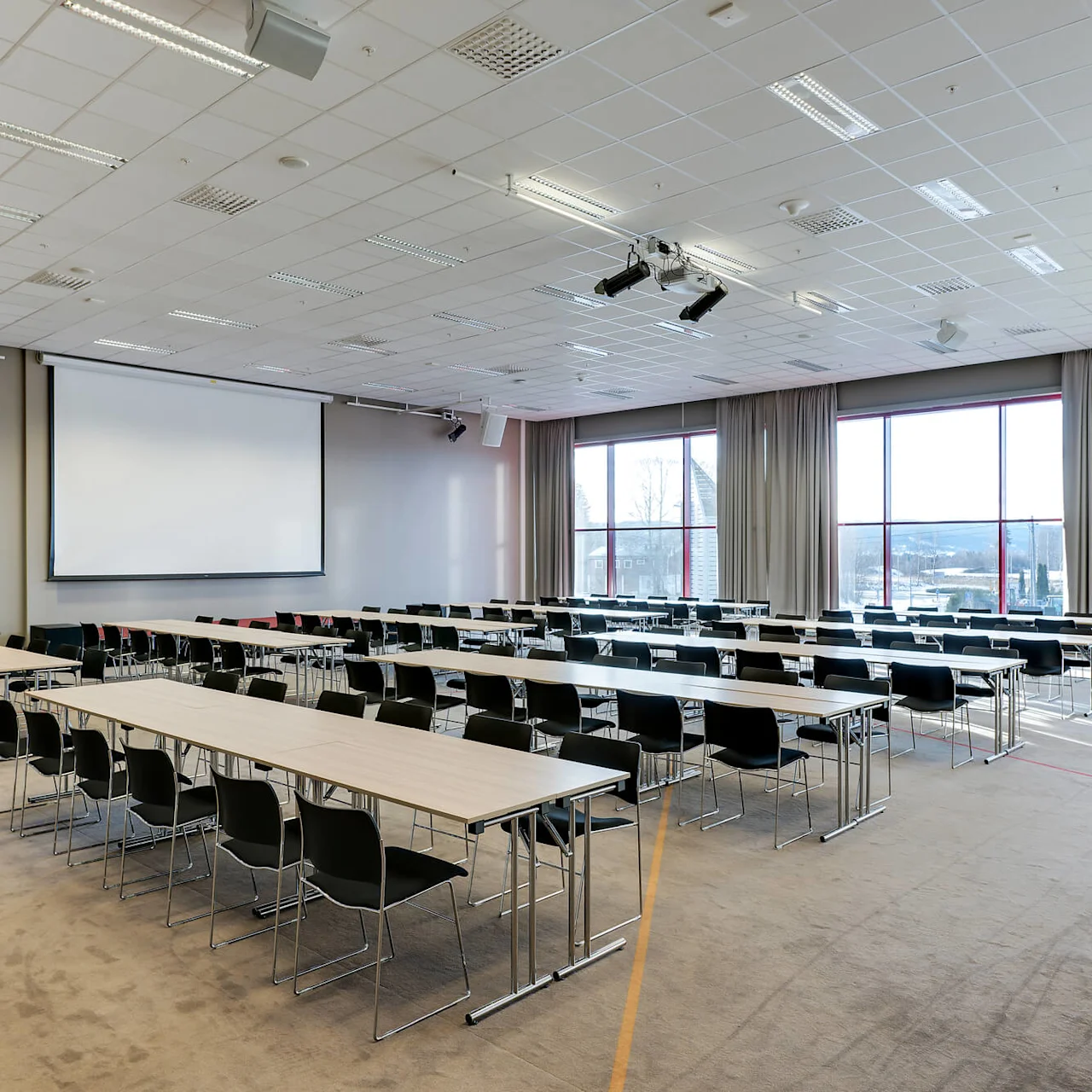 Ett konferensrum med tomma rader av bord och stolar vänder sig mot en stor projektorduk. Solljus strömmar genom de höga fönstren och belyser den moderna, rymliga miljön.