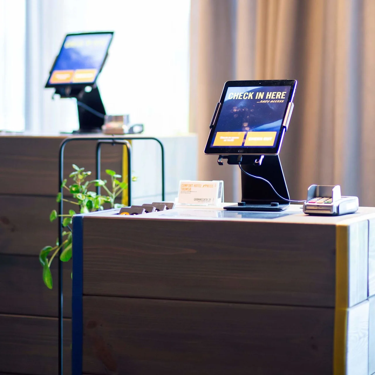 Tablet on stand displaying "CHECK IN HERE - EASY ACCESS," next to a card reader, on a wooden counter in a lobby. Background shows another tablet and plant. Nearby sign reads "COMFORT HOTEL XPRESS TROMSØ."