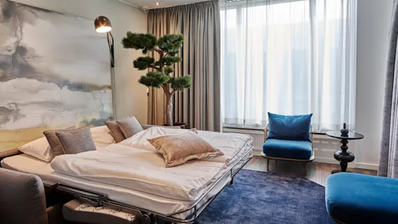 Sengen er pent redd opp med puter, plassert nær et stort maleri under en vegglampe. Omgitt av et lite tre, gardiner, en blå stol og bord i et koselig rom.