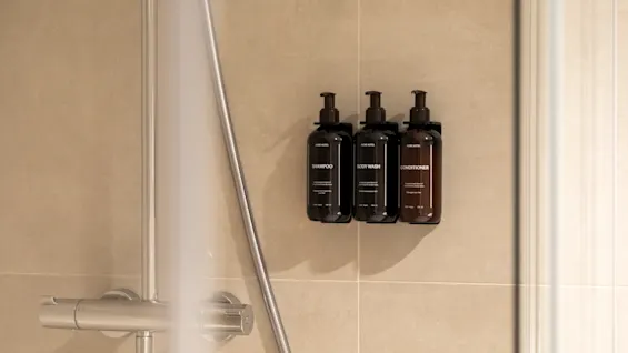 Three dark dispenser bottles labeled SHAMPOO, BODY WASH, and CONDITIONER mounted on a tiled shower wall.