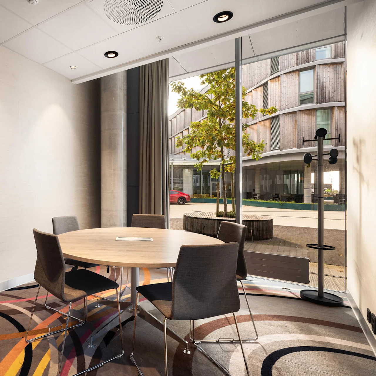 Round table with four chairs sits on a colorful carpet in a modern office. Large windows reveal a view of a tree and a red car outside.
