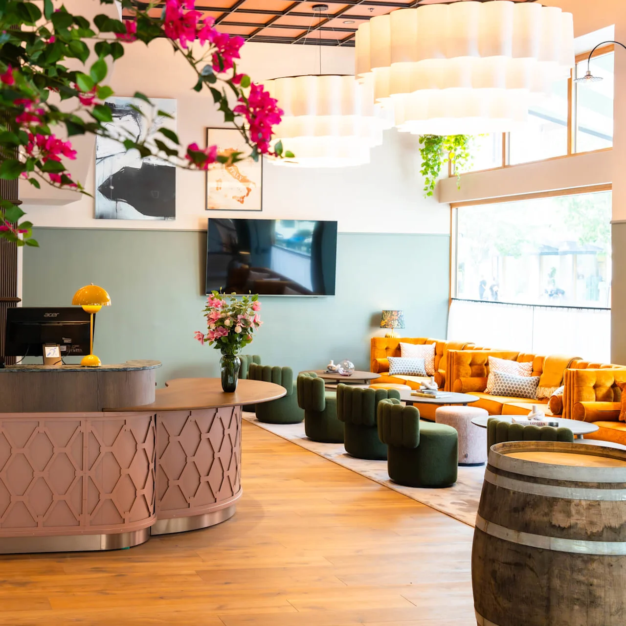 Reception desk with a person behind it, typing on a computer. Bright pink flowers hang in the foreground. Cozy seating area with yellow sofas and green chairs in the background.