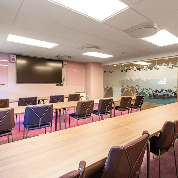 A conference room features two tables forming an L-shape with chairs, a large wall-mounted screen, and a podium. Carpeted flooring, frosted glass wall, and wooden doors set the modern ambiance.